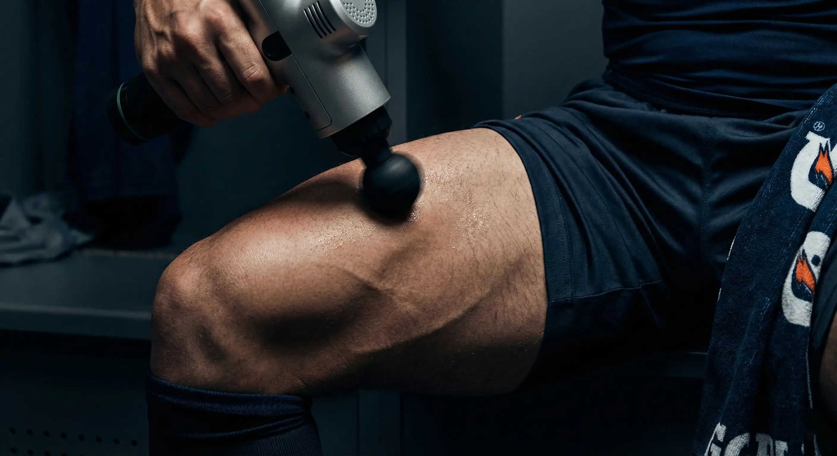 Masajeador de percusión profesional aplicado en cuádriceps de un futbolista veterano para aliviar la fatiga muscular tras el partido.