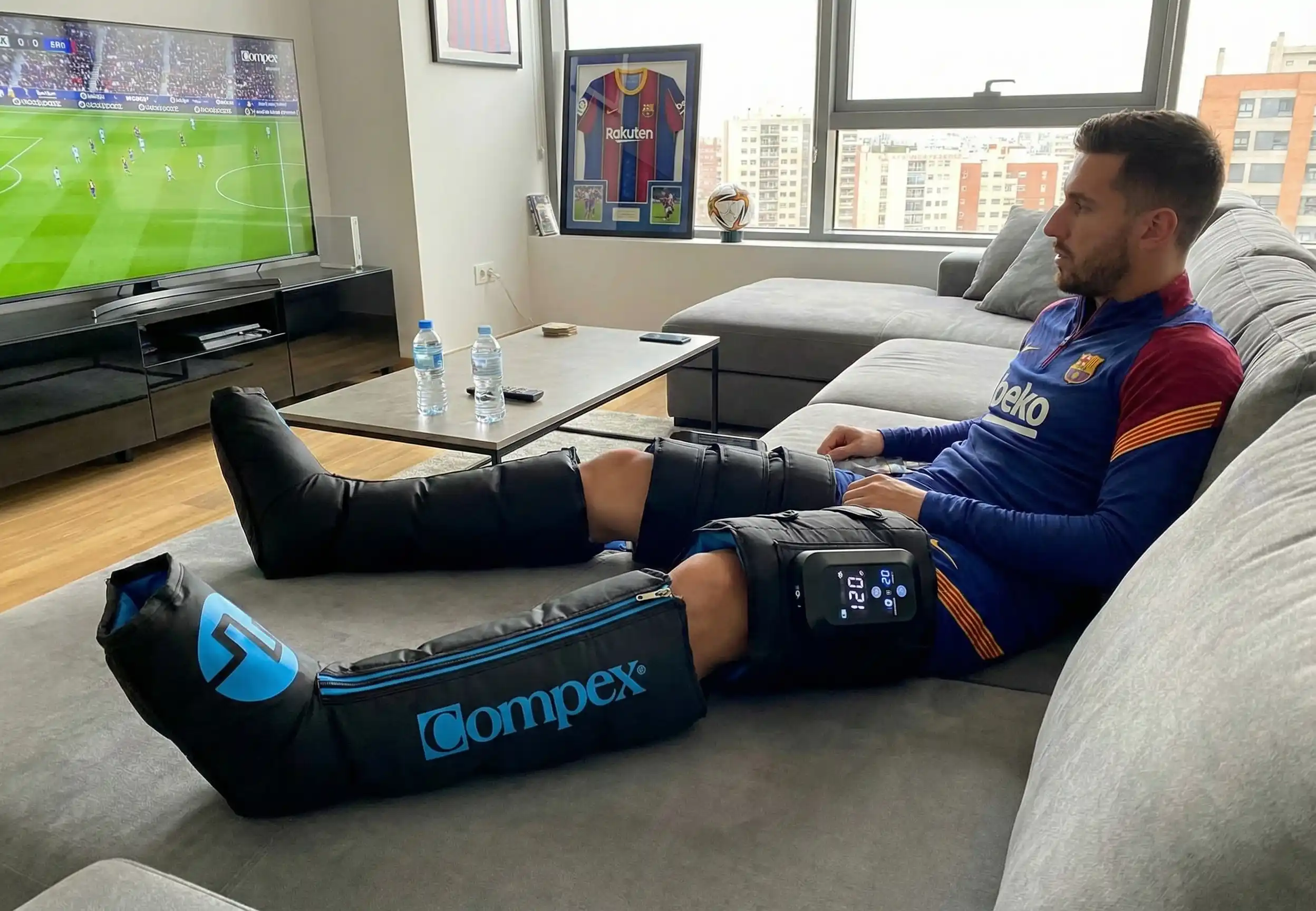 Futbolista profesional utilizando botas de presoterapia inalámbricas de última generación en el sofá de su casa, mostrando el panel de control digital y el inflado de cámaras