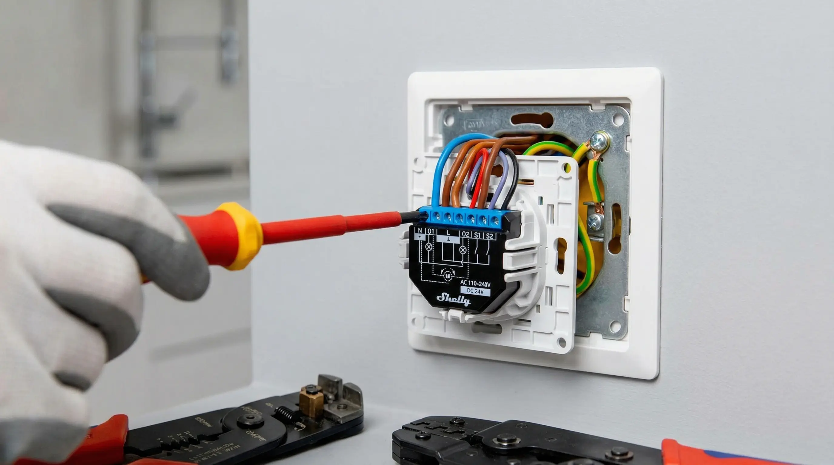 Técnico profesional instalando un módulo Shelly Plus 2PM en una caja de pared española. Se muestran los cables con colores estándar: azul para neutro, marrón para fase y verde-amarillo para toma de tierra. Conexiones ordenadas y herramientas profesionales visibles en un entorno doméstico moderno.