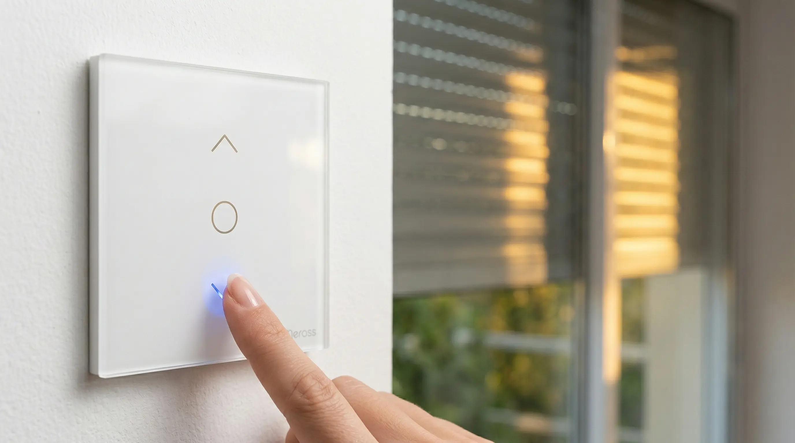Primer plano de un interruptor táctil Meross en una pared blanca. Se aprecia el logo de Meross y un indicador LED azul sutil. Un dedo toca el panel mientras de fondo una persiana de aluminio baja suavemente filtrando la luz del atardecer. Estilo minimalista y enfoque nítido.