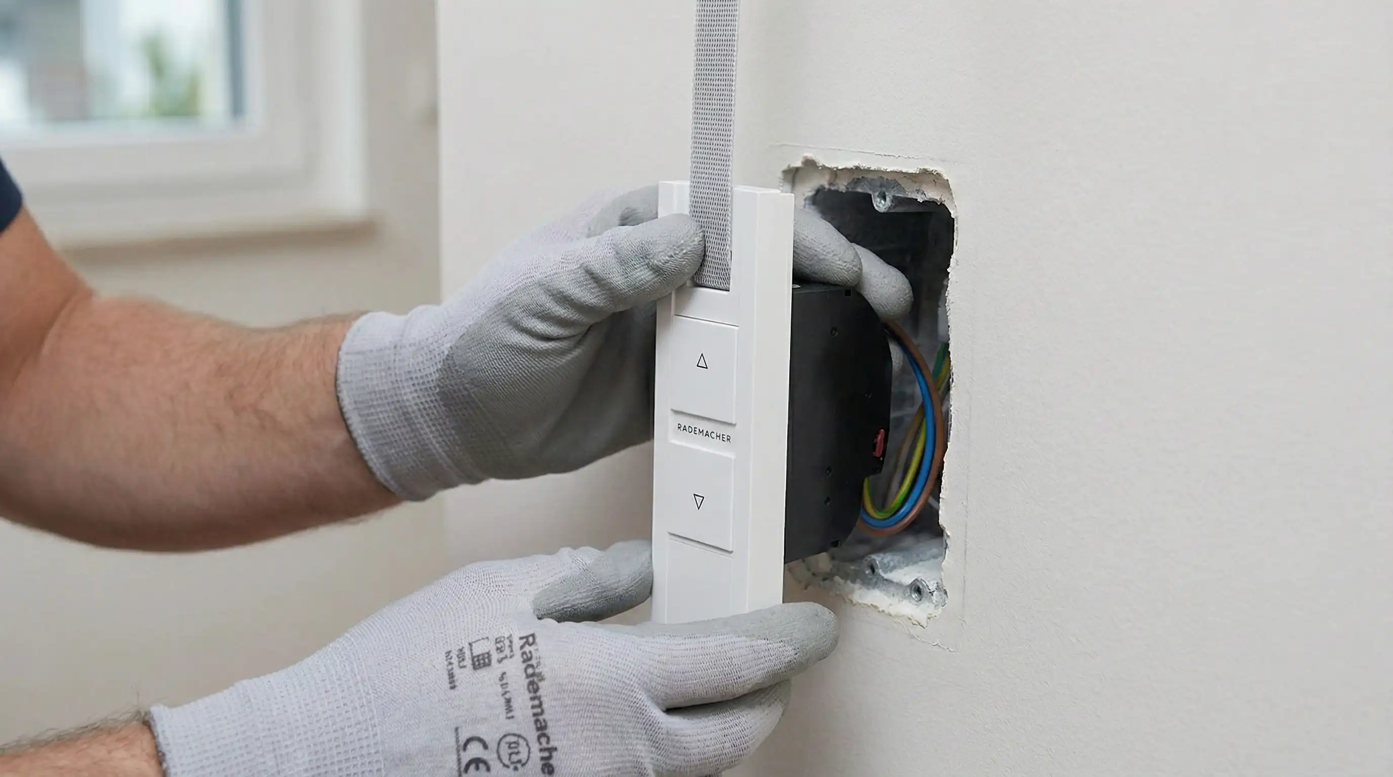 Primer plano de manos de un técnico encajando un enrollador eléctrico blanco en un hueco de pared estándar. Se ve la cinta de la persiana entrando limpia y recta por la ranura superior del dispositivo. Luz clara, enfoque en el mecanismo.